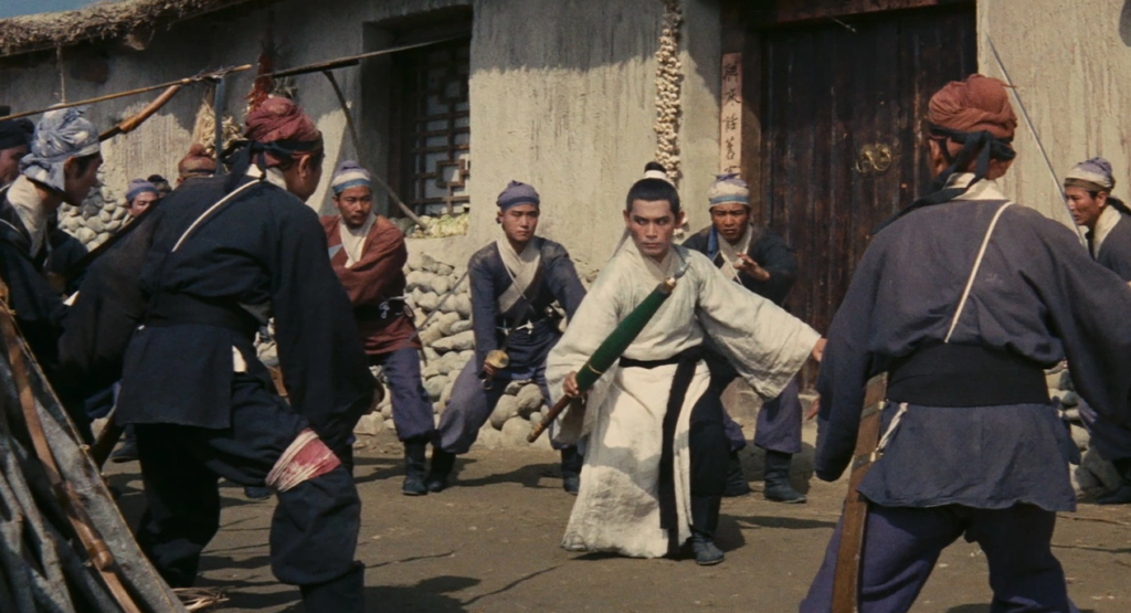 A martial artist stands in front of an inn. He is surrounded by men bearing swords and spears.