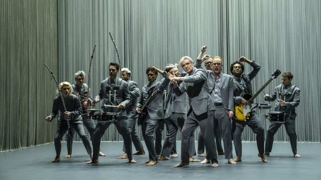 A group of barefoot musicians are all wearing grey suits and playing instruments