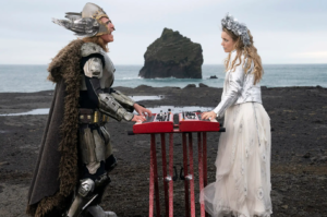 A man in stylized armor and a woman in a stylized crown play keyboards on a beach