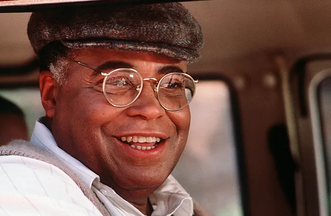 A man in glasses and a flat cap sits in a car and smiles