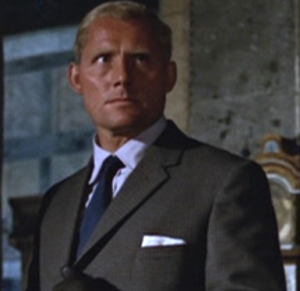 An Irish spy with platinum blond hair is in a gray suit with blue tie. He is looking menacingly off the screen to stage left.