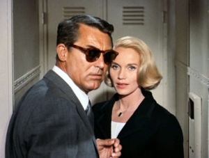 A figure in a grey flannel suit and dark sunglasses stands in a train hallway next to a blonde in a black jacket and white dress