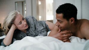 Two people in underwear lay on a white duvet. The woman is leaning on her hand and seems pensive. The man is touching his face and grinning