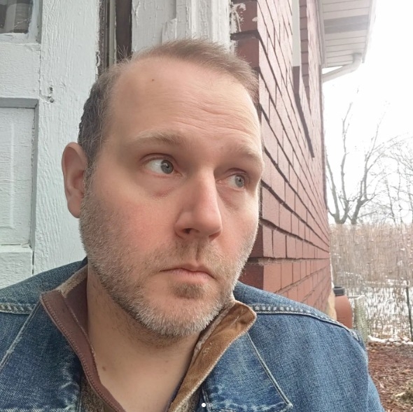 Matt sits in a doorway of a brick house and looks off-camera. He is wearing a denim jacket and tan sweater.