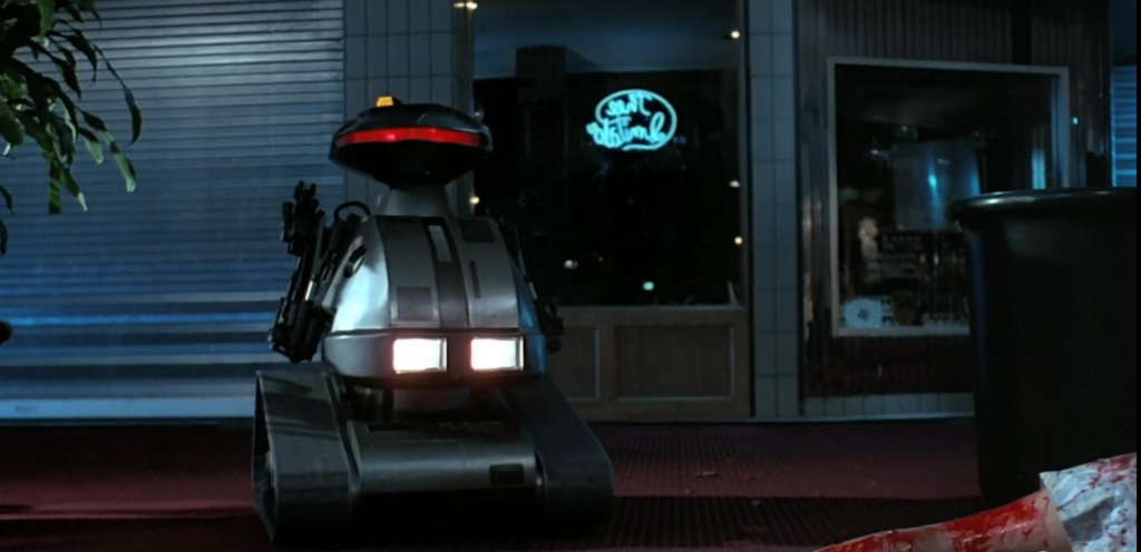 A grey robot with tank treads and a red light on its visor rolls past the corpse of a woman. This is in the food court of a shopping mall