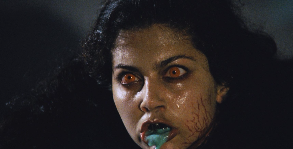 A possessed woman with long black hair has red pupils and has green slime dripping from her mouth