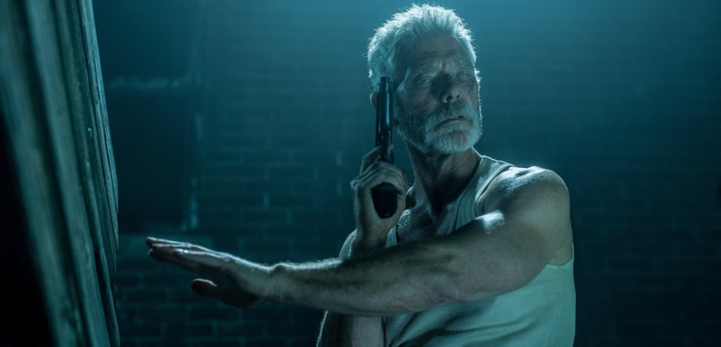 A blind man with short grey hair and a bear stands in a tank top in a basement. He holds a pistol and is reaching out for a wooden stairwell with his other hand.
