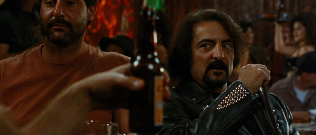 A man with long black hair and a goatee looks across a barroom table toward someone holding a beer bottle.