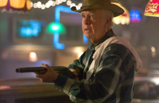A senior bartender wears a cowboy hat and has a dishtowel on his shoulder. He holds a shotgun