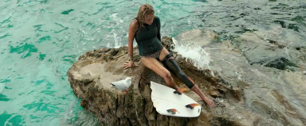 A surfer sits on a rock with a seagull. The surfer has a tourniquet around her leg because of a shark attack