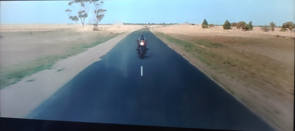 Screenshot from Mad Max (1979). A biker flees on a highway, pursued by a policeman in a black sports car that is just out of sight.