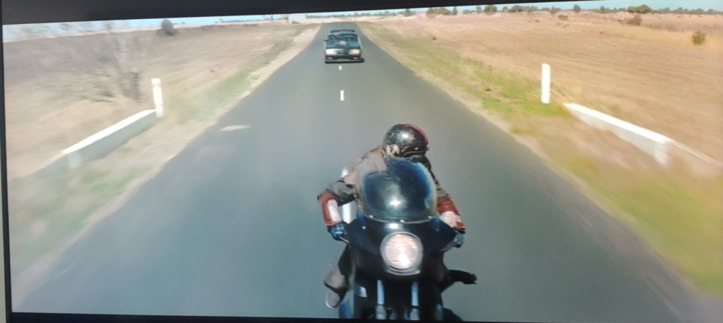 Screenshot from Mad Max (1979). A biker is still fleeing from a police car, and the car is much closer to the biker, who is looking over their shoulder at the car.