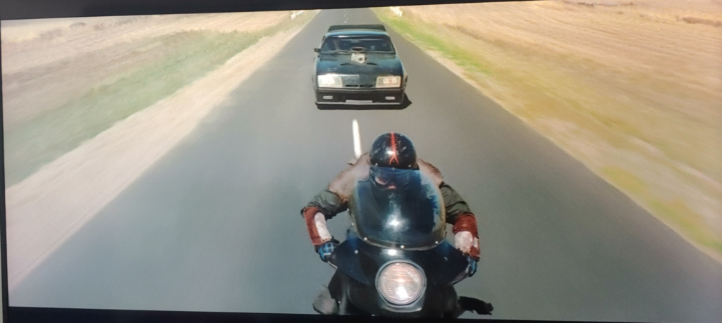 Screenshot from Mad Max (1979). The police car has caught up to the fleeing biker and is nearly on the biker's back tire.