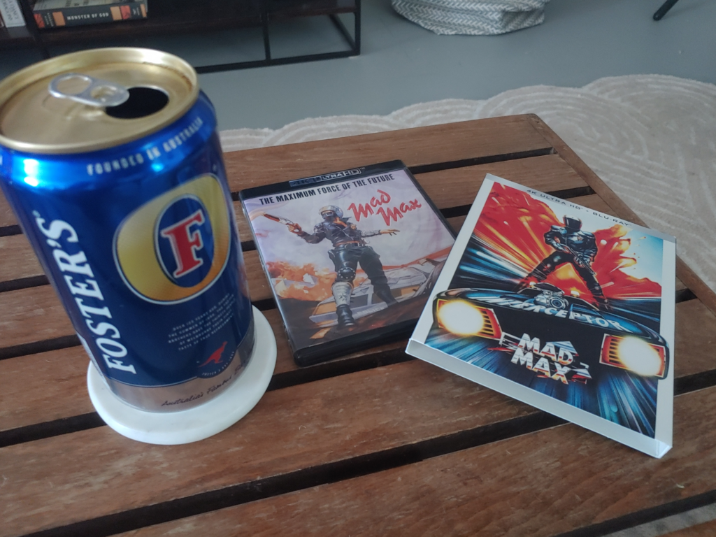 A can of Foster's beer sits on a table next to the case for a 4k version of the film Mad Max