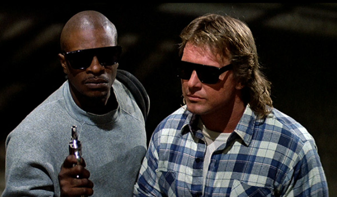 Two men in dark sunglasses hold pistols and look determined