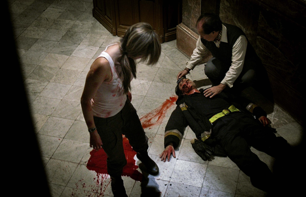 A news reporter in a white tank top looks down at a doctor holding the body of a paramedic.