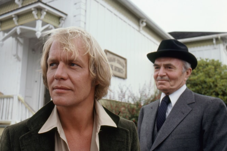 A blonde man looks worried and stands in front of an antique shop. An older man stands behind him in a suit and hat and looks at the front man