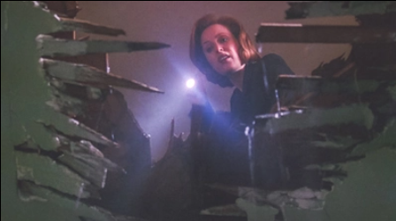 FBI Agent Scully uses a flashlight to peer down a torn hole in the floor of a building