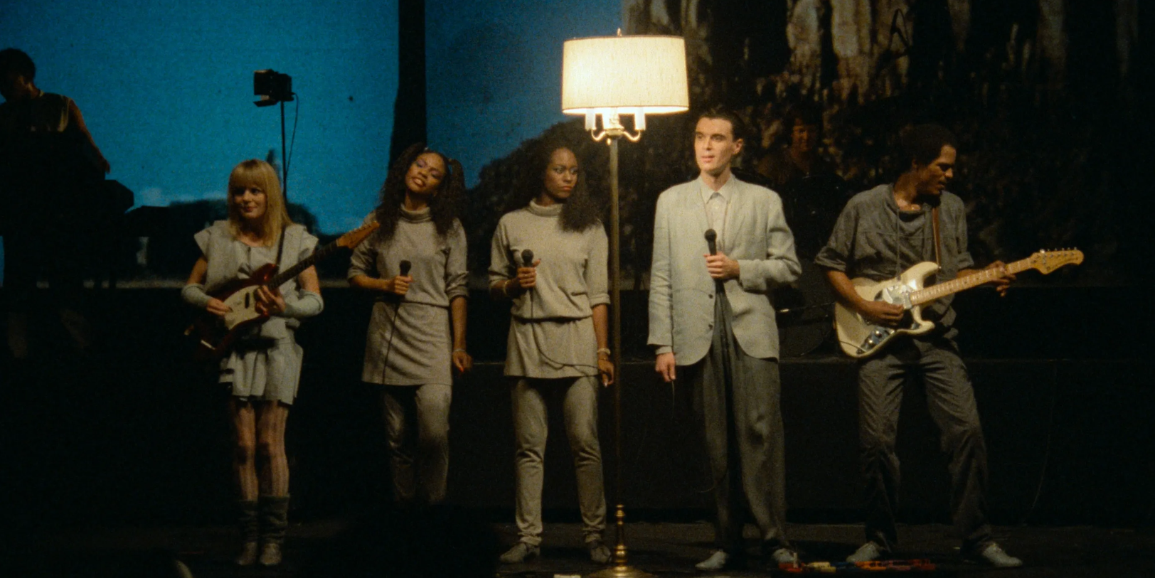 The members of Talking Heads perform on a stage with a large floor lamp behind them.